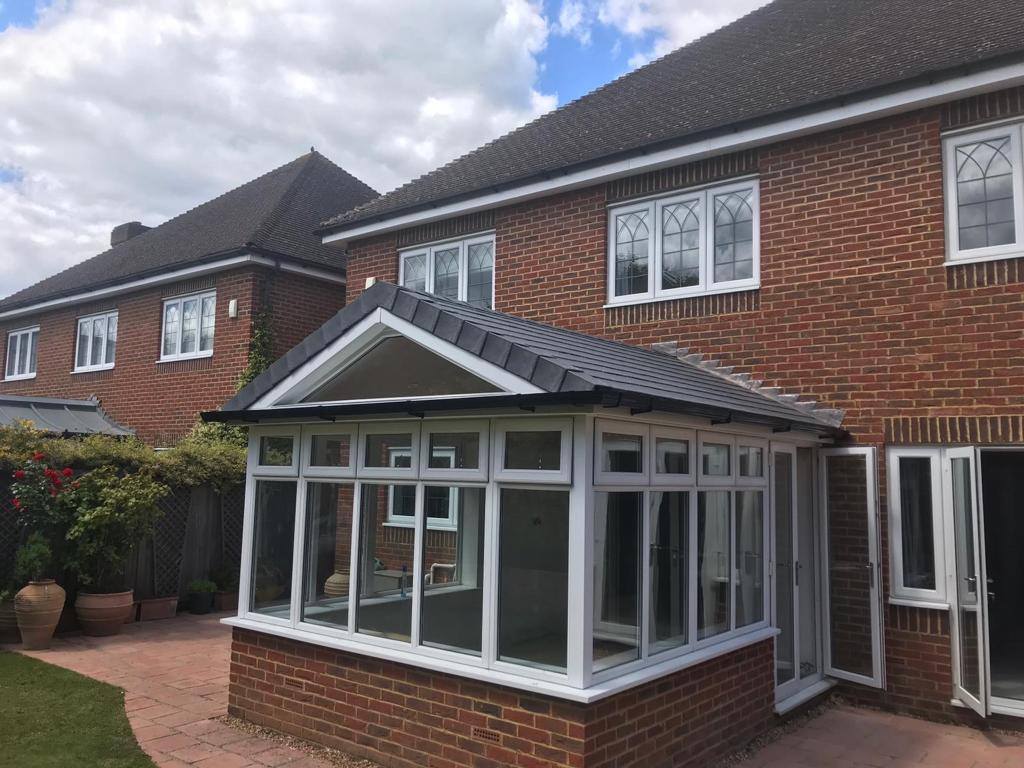 A newly installed conservatory with a classic white frame and glass panelling, attached to a red brick house with a hipped roof. The conservatory features a lean-to roof style, complementing the traditional architecture of the house, set in a garden with a brick patio and greenery.