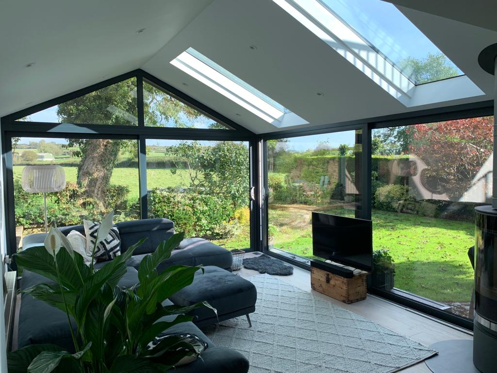 A stylish interior of a modern conservatory with a vaulted ceiling, featuring skylights, black framed glass doors, and panoramic windows offering views of a verdant garden. The space is furnished with a luxurious dark sectional sofa, a large flat-screen TV, and complemented by indoor plants and a chic floor lamp.
