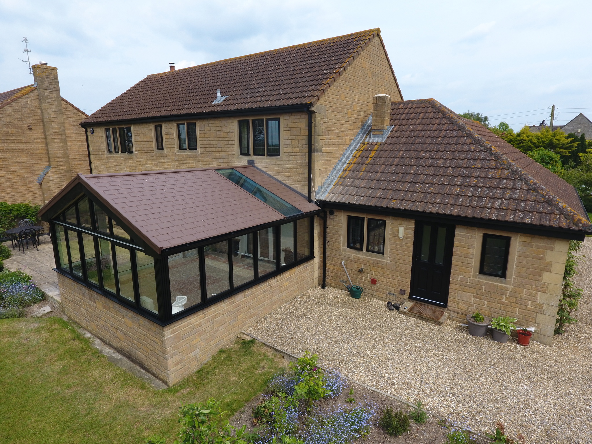 Aerial view of a stone-built house featuring a large, brown-framed conservatory with a gabled roof and glass walls. The conservatory is set in a well-kept garden with a gravel path and is complemented by the surrounding greenery and floral beds