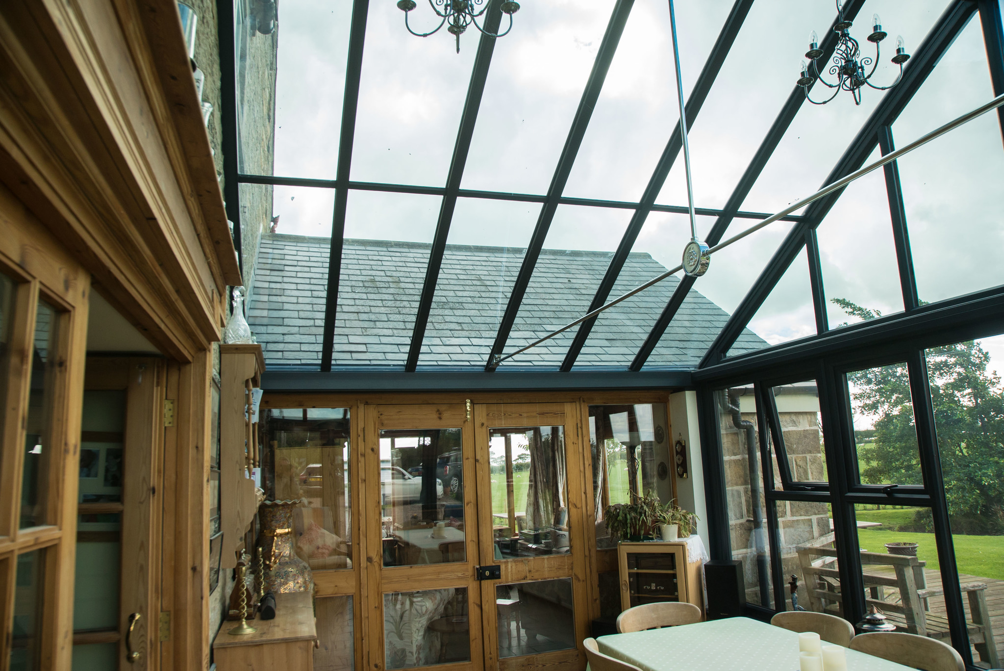 Interior view of a rustic-themed gable conservatory with a high-pitched ceiling, featuring exposed wooden beams, classic chandelier lighting, and a cozy seating arrangement, all bathed in the warm glow of natural light filtering through the expansive glass facade
