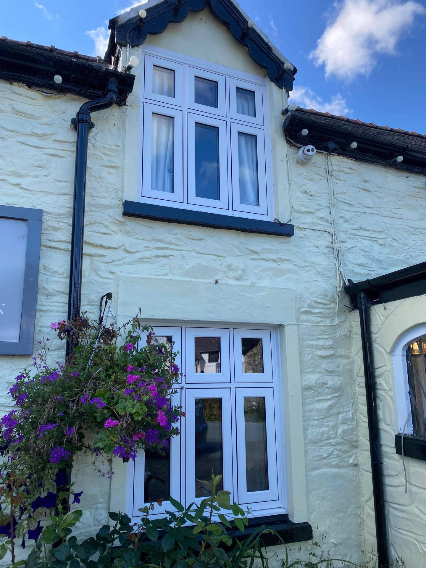 raditional white cottage with a textured finish, adorned with dark-trimmed uPVC casement windows. The upper window sits within a gabled dormer, while the lower window is framed by cascading purple flowers from a hanging basket, adding a vibrant splash of color to the quaint facade.