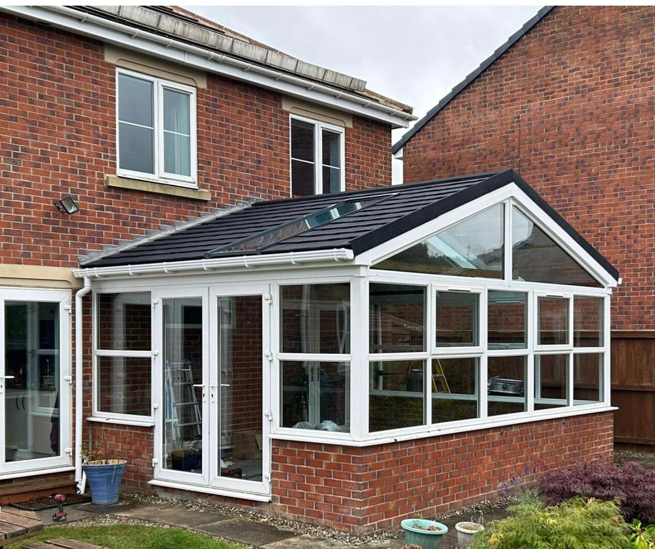 A lean-to conservatory with a gabled front and a black tiled roof, attached to a red brick house. The conservatory has white frames, large windows, and French doors, nestled in a garden with potted plants and a neatly trimmed lawn