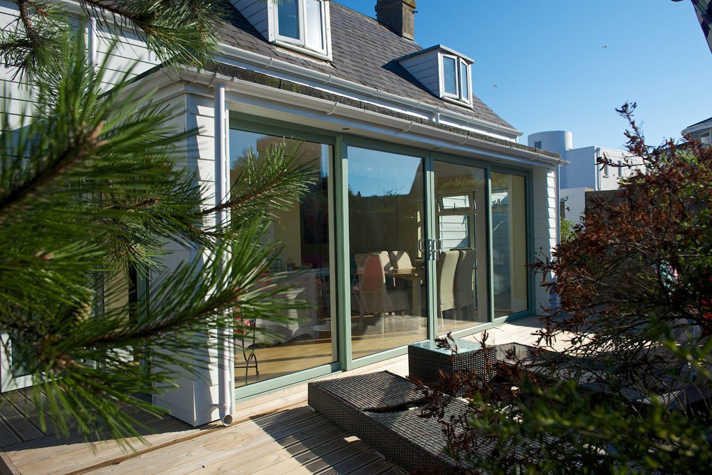 A modern conservatory with large glass doors opening onto a wooden deck. The conservatory is attached to a white house with a dark grey roof, complemented by the clear blue sky in the background. Outdoor furniture is partially visible inside, suggesting a seamless indoor-outdoor living space. Greenery frames the scene, adding a touch of nature to the home extension.