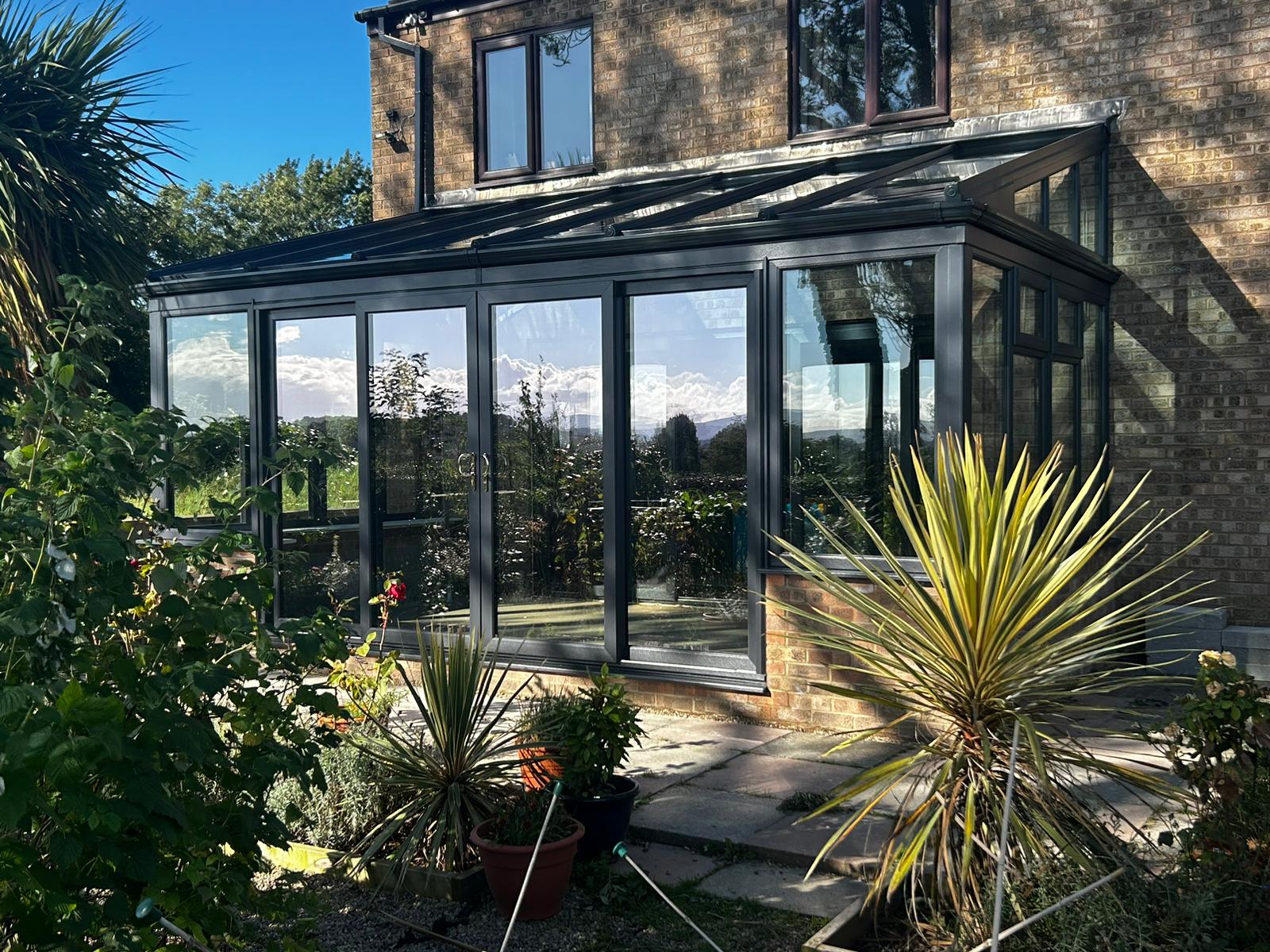 an elegant lean-to conservatory with a black frame and glass panels, situated in a lush garden