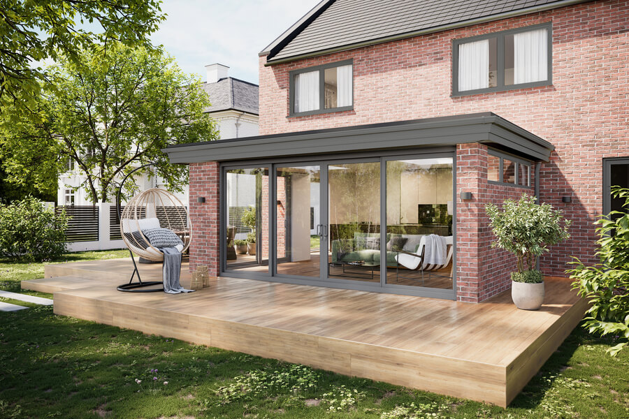 A grey sunroom extension with a glass roof attached to the back of a red brick house. The sunroom has large windows and a double door, and it's built on a wooden deck. The interior shows a dining table and chairs, and a plant sits on a small table. On the deck outside, there are two outdoor chairs and a small table.