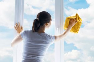 image of woman cleaning a upvc window with a yellow cloth, angled to see bright blue sky and clouds outside